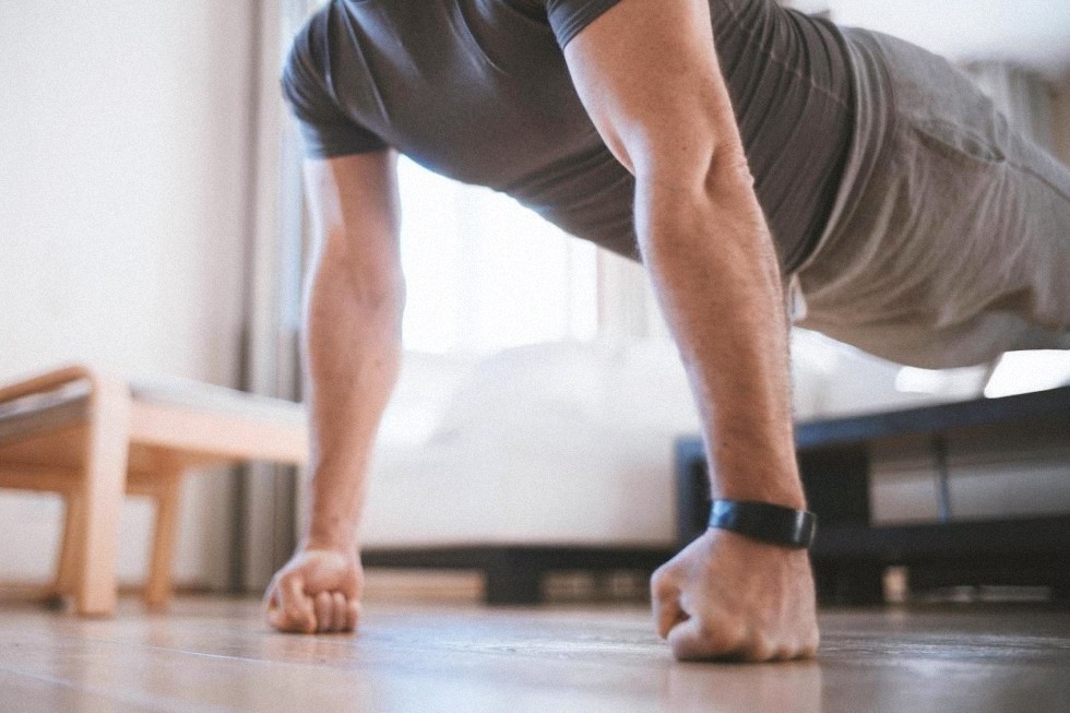 Persona haciendo flexiones apoyada en los nudillos en un espacio interior, vestida con una camiseta de manga corta y con un pulsera de actividad, con muebles visibles al fondo, realizando ejercicios de calistenia militar para desarrollar fuerza y resistencia.