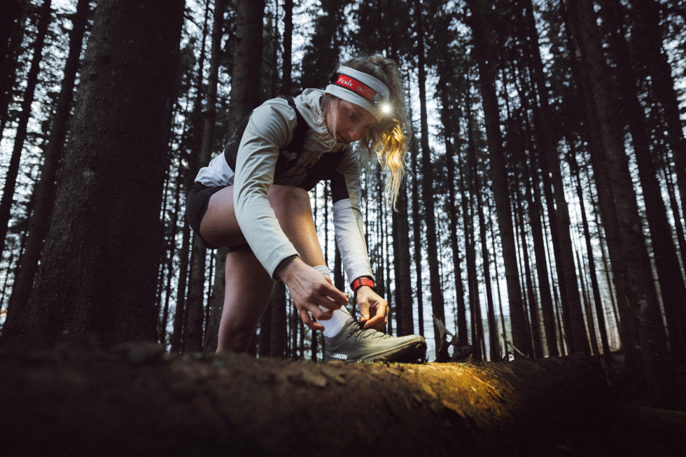 Corredora de trail running se ata los cordones con el pie apoyado en un tronco, mientras una linterna frontal Fenix ilumina su zapatilla en un bosque con poca luz.