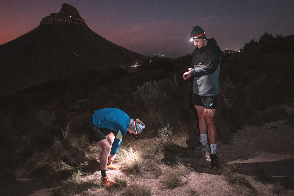 Dos corredores de trail running con linternas frontales Fenix en un sendero de montaña de noche; uno se ajusta el calzado y el otro consulta su reloj.