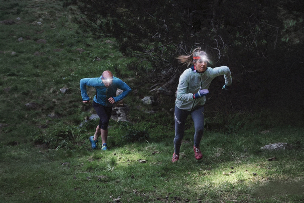 Dos corredores de trail running suben una ladera boscosa con linternas frontales Fenix que iluminan el terreno con poca luz.