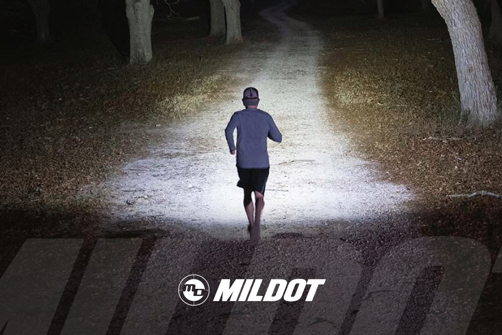 Corredor de trail running visto de espaldas en un sendero de bosque de noche, iluminado por una linterna frontal, con el logotipo de MILDOT en la parte inferior.