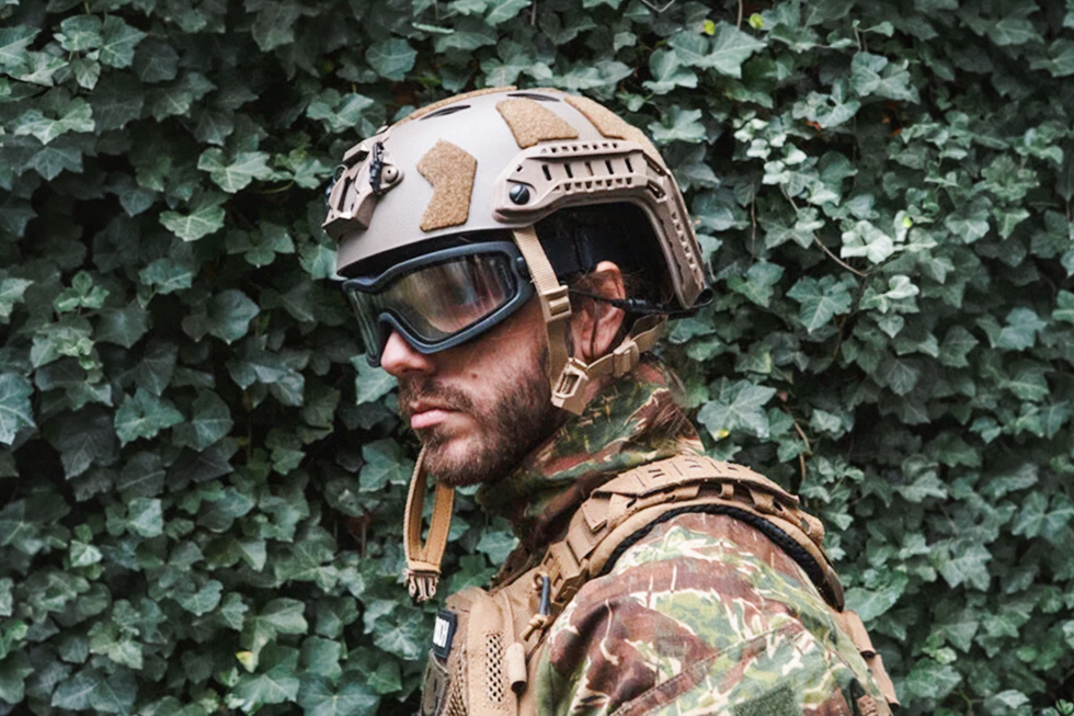 Jugador de airsoft con gafas de protección cerradas, casco táctico y uniforme de camuflaje frente a una pared de hiedra.