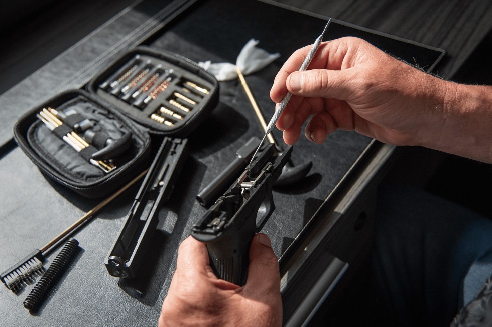 Persona realizando el mantenimiento y la limpieza interna de una réplica de pistola de airsoft desmontada sobre una mesa, utilizando herramientas de precisión y un kit de limpieza