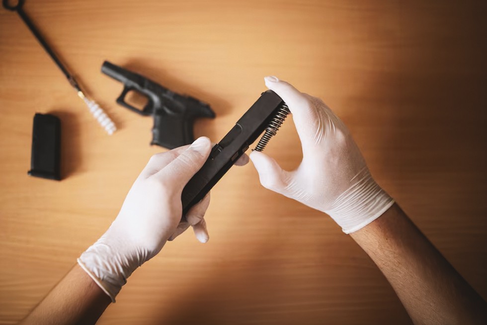 Persona con guantes realizando el desmontaje y mantenimiento de la corredera de una réplica de pistola de airsoft, con el cargador y un kit de limpieza sobre una mesa de trabajo