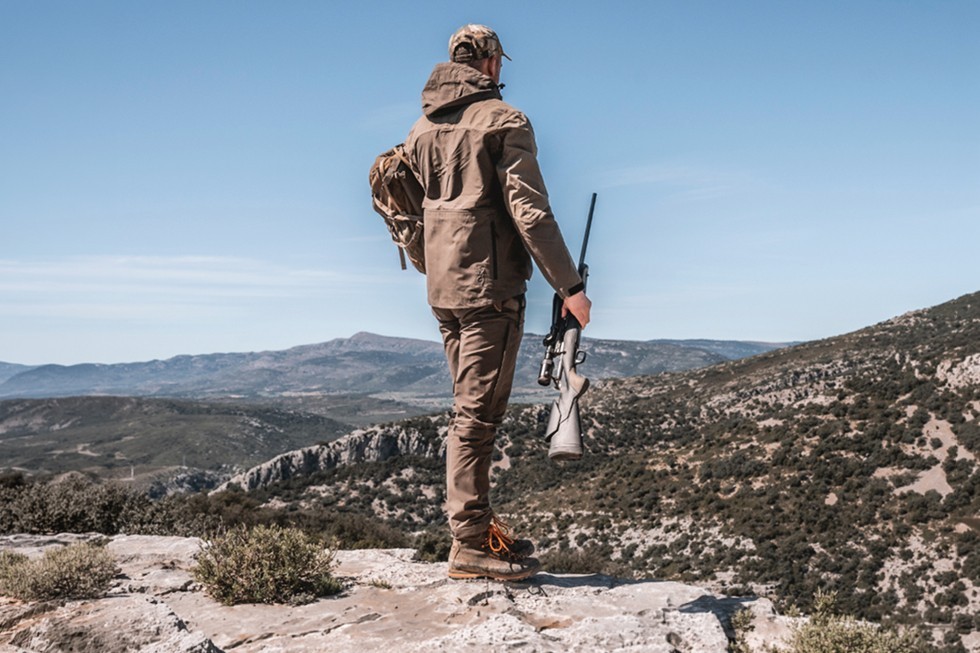 Cazador con ropa de caza marrón y rifle recorriendo un terreno montañoso.