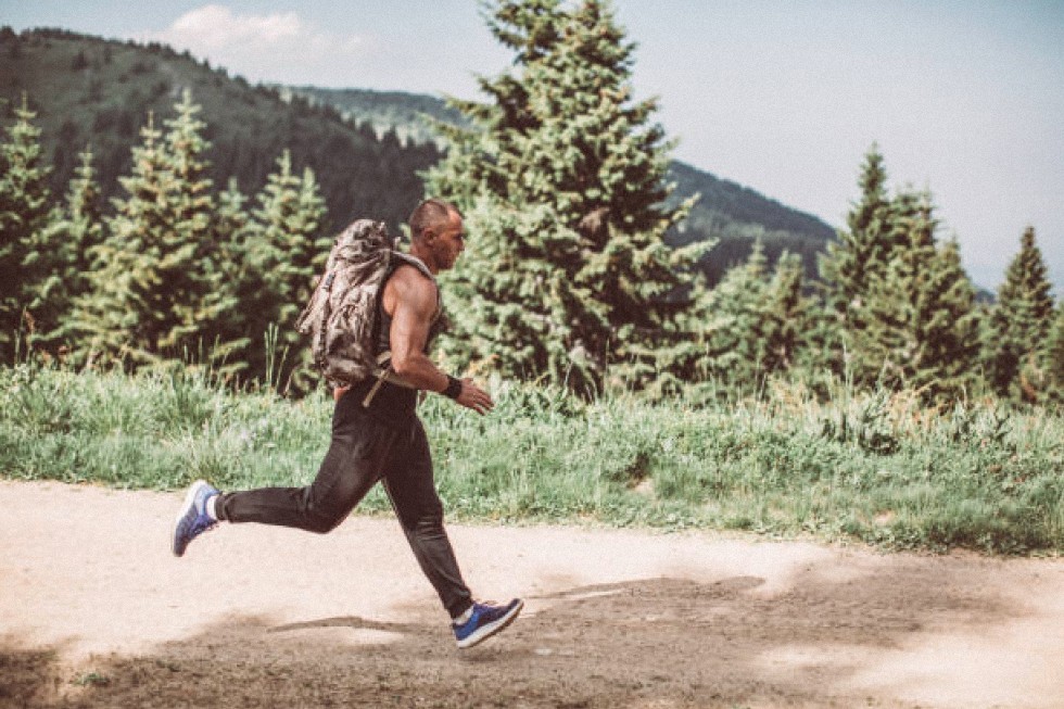 Una persona que practica calistenia militar, vestida con una camiseta sin mangas y una mochila, corre por un sendero de tierra que atraviesa una zona boscosa con pinos y una montaña al fondo.