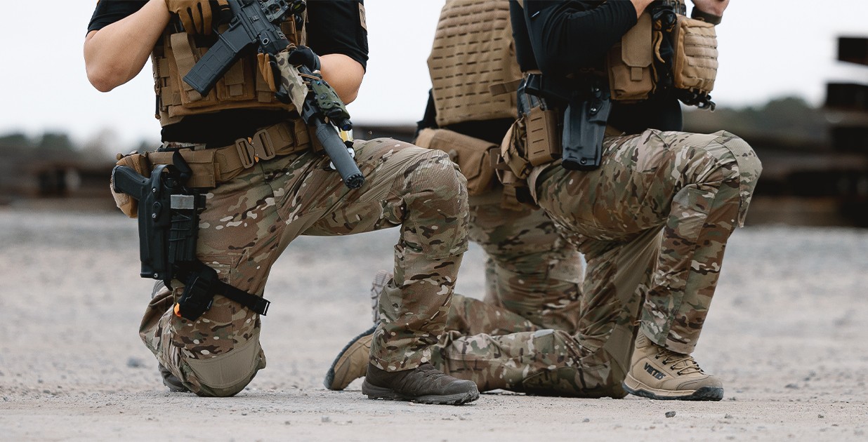 Dos operadores tácticos arrodillados con pantalones Condor Paladin en camuflaje multicam, equipados con chalecos tácticos, cinturones modulares y armas de entrenamiento en un entorno al aire libre.